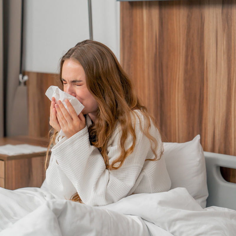 Vrouw in badjas niest in een tissue terwijl ze in bed zit, mogelijk door huisstofmijtallergie.